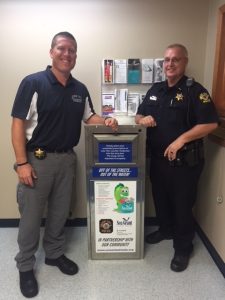 Two police officers stand next to a medicine collection box with IISG branding