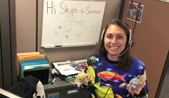 Katie O'Reilly sits at her desk in a fish shirt, holding fish toys, with "Hi, Skype a Scientist!" written on a whiteboard along with a hand-drawn fish.