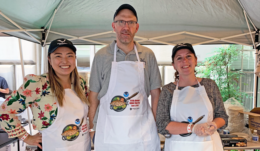 two women and a man stand under a tent wearing Illinois-Indiana Sea Grant hats and white NOAA Fish Fry aprons