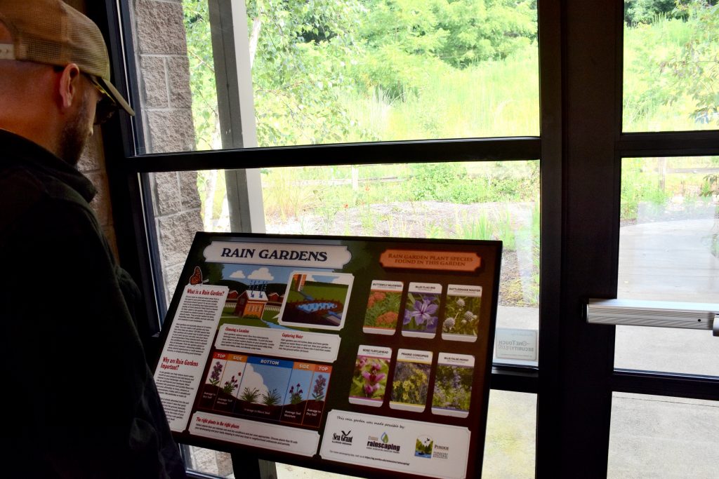 man rading sign labeled "Rain Gardens" with info about plant species