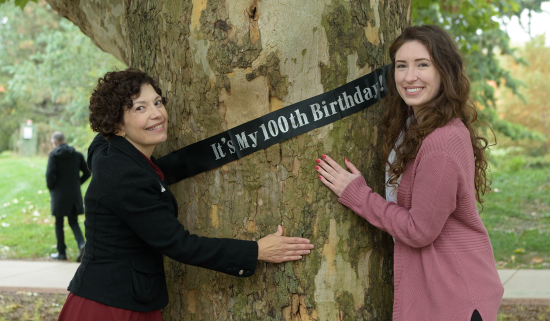 two women hug a tree with a banner that reads "it's my 100th birthday!"