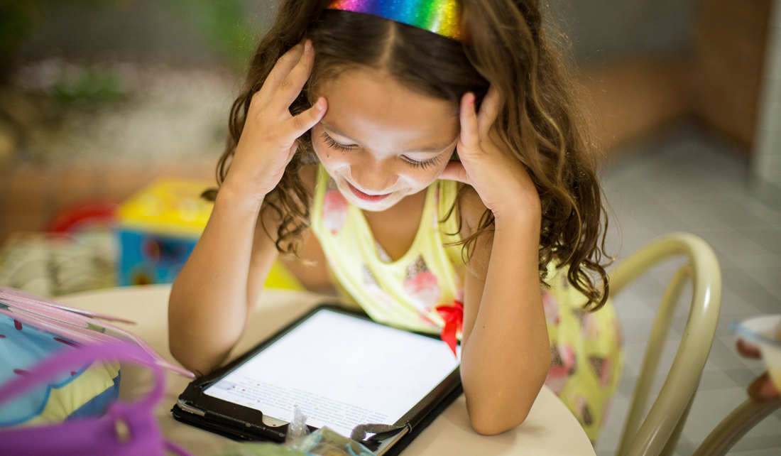 Young girl sits at a table and smiles down at an electronic tablet, backpack sitting beside her