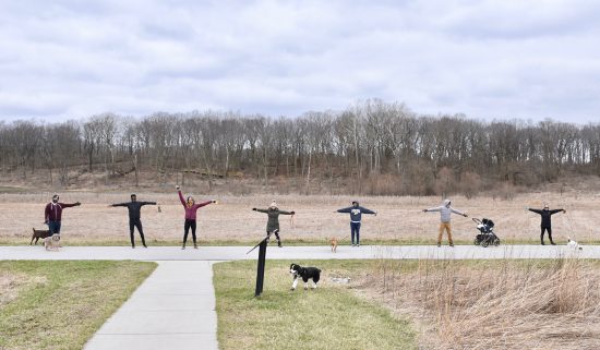 several people stand on a park path with their arms out, at least 6 feet apart from the next person