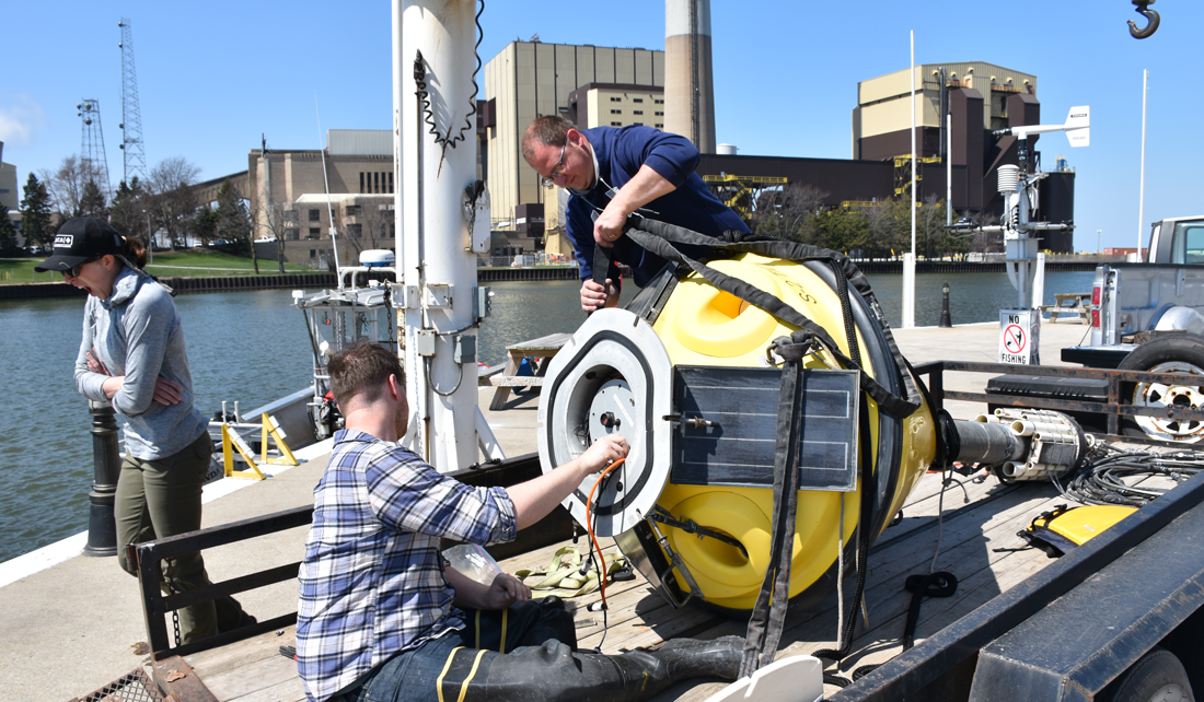 Two people work on a large, yellow buoy on the back of a trailer parked next to Lake Michigan