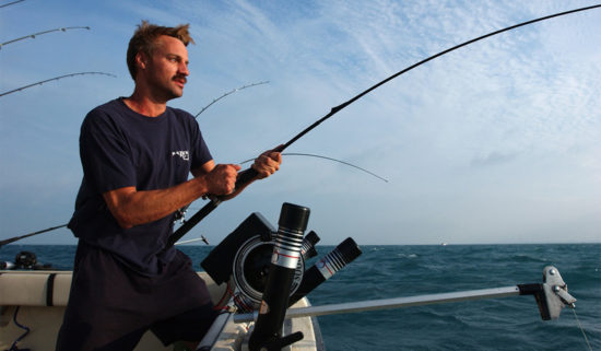 man with mustache stands near edge of boat holding fishing pole