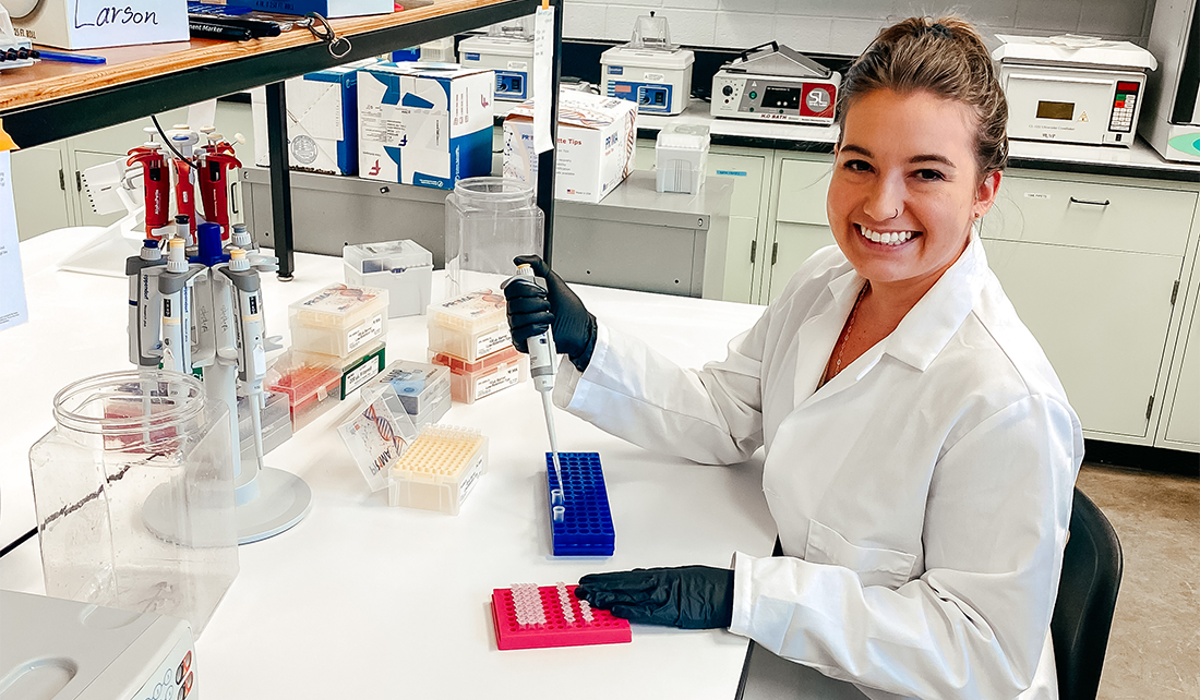woman sits at lab bench with lab coat and gloves on, using DNA extracting and sequencing techniques