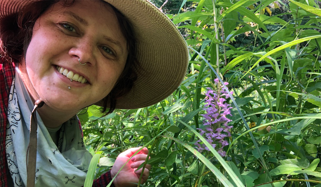 woman wearing bandana and safari hat stands in tall grasses and plants, and smiles next to a rare pink orchid