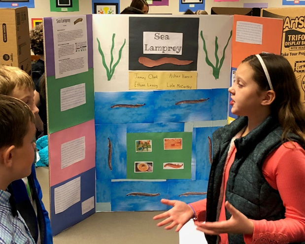girl stands in front of sea lamprey science poster, giving a presentation to other students