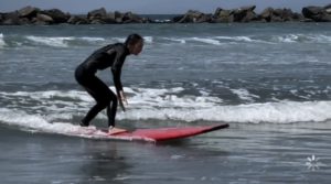 Wavy water. Person in black wet suit on red surfboard. Rocky shore in background.