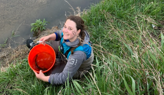 Smiling person in lower left, holding an orange bucket. This person is sitting on a grassy stream bank. The stream is reflecting the sky and there is a net enclosure in the middle of the stream.