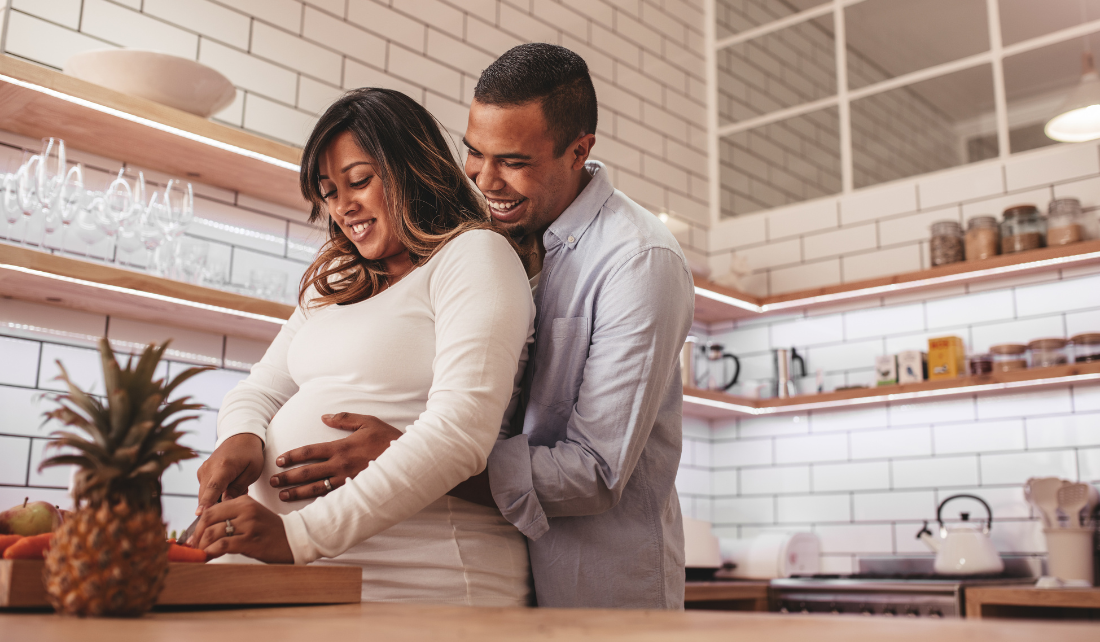 Image of a man and woman cooking dinner in the kitchen. The man holds the woman's pregnant belly as she cooks.