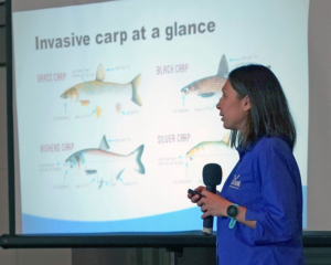 Katie O'Reilly stands with microphone in hand, in front of a projector screen showing a graphic titled "invasive carp at a glance"