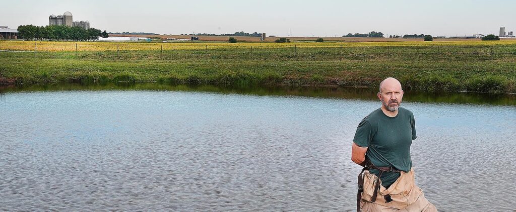 Tomas Hook poses in front of pond with waders on and farmland in the background