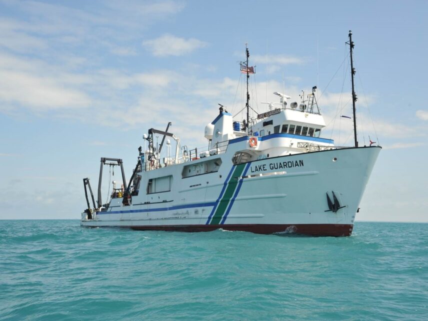 Research vessel Lake Guardian underway on calm turquoise water beneath a blue sky with scattered clouds. The white ship, equipped with cranes and scientific equipment, is viewed from the front starboard side.